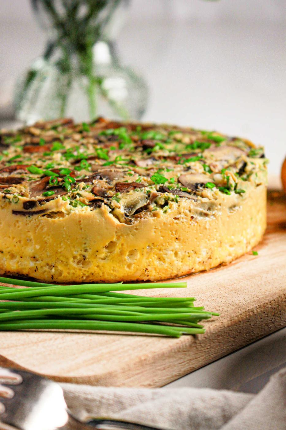 A thick vegetable frittata, inspired by an Instant Pot quiche recipe, is topped with herbs and sits on a wooden board next to fresh chives.