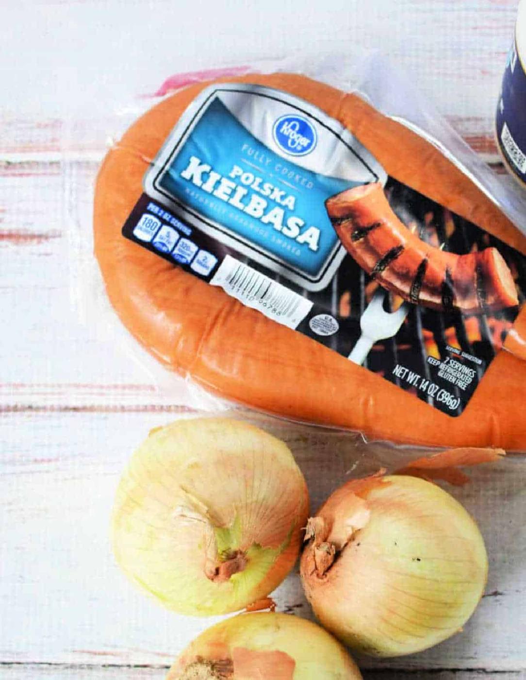 Packaged Polska kielbasa sausage next to three yellow onions on a light wooden surface—perfect ingredients for making instant pot red beans and rice.