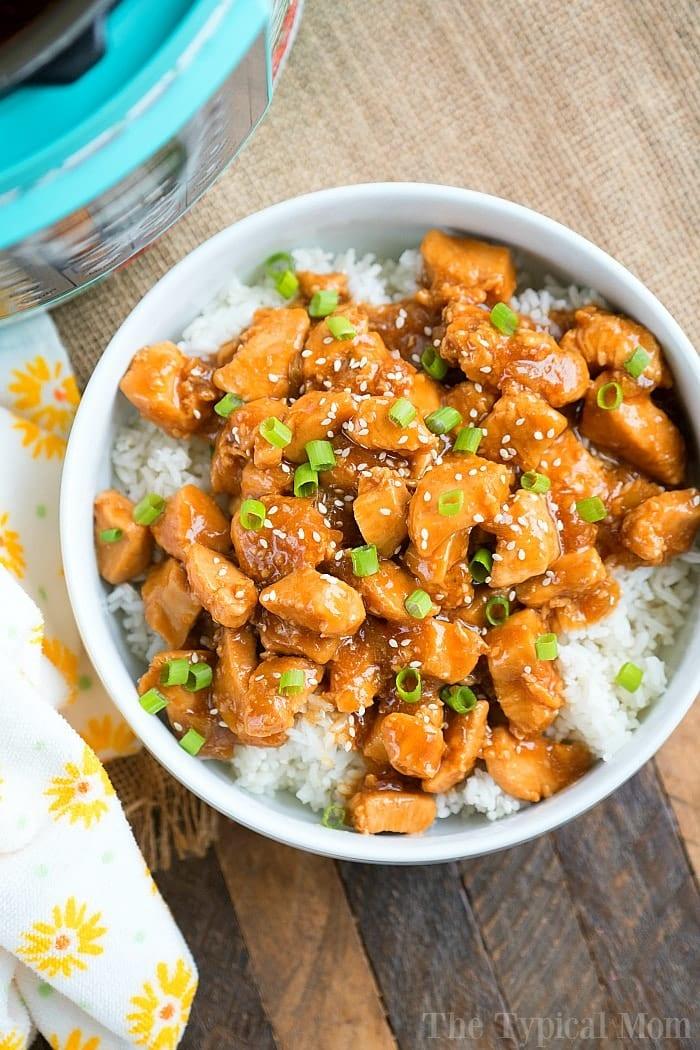 pressure cooker sesame chicken, perfectly glazed and sprinkled with green onions, rests atop a bed of rice on a wooden surface. A floral napkin adds an elegant touch to the setting.