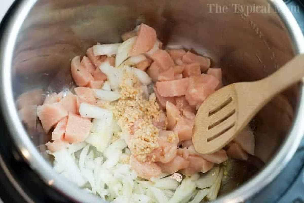 Chopped raw chicken, onions, and minced garlic in a pot with a wooden spoon—perfect for starting your instant pot white chicken chili.