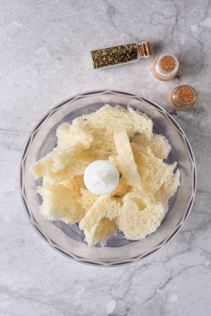 A food processor bowl contains torn pieces of bread, soon to become Italian Panko Bread Crumbs. Next to it are two small cork-stoppered bottles with spices and herbs, along with a small container of ground spice, all resting on a speckled gray countertop.