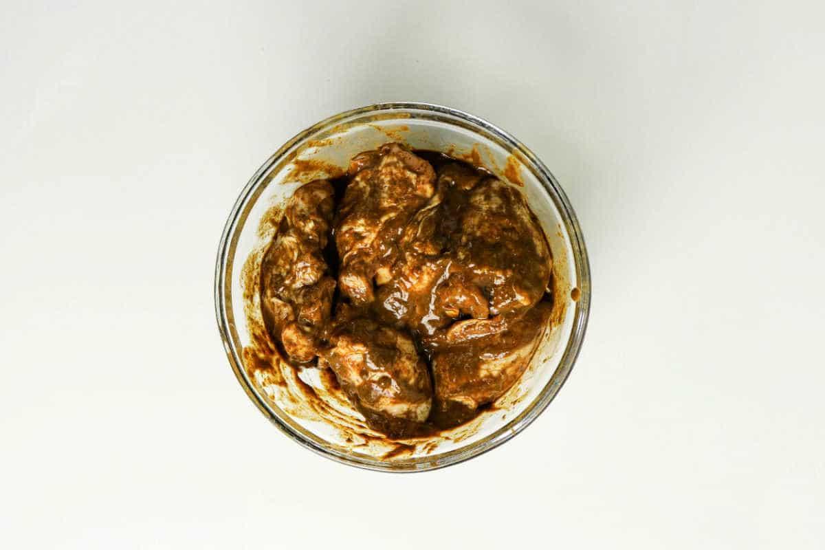 Glass bowl with pieces of meat coated in a thick brown jerk chicken marinade, ready for the oven, placed on a white surface—a perfect start to your favorite jerk chicken recipe.