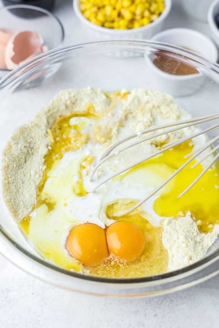 A mixing bowl with flour, two cracked eggs, and milk being whisked; corn and other ingredients in the background ready to create a delicious jiffy cornbread in a cast iron skillet.