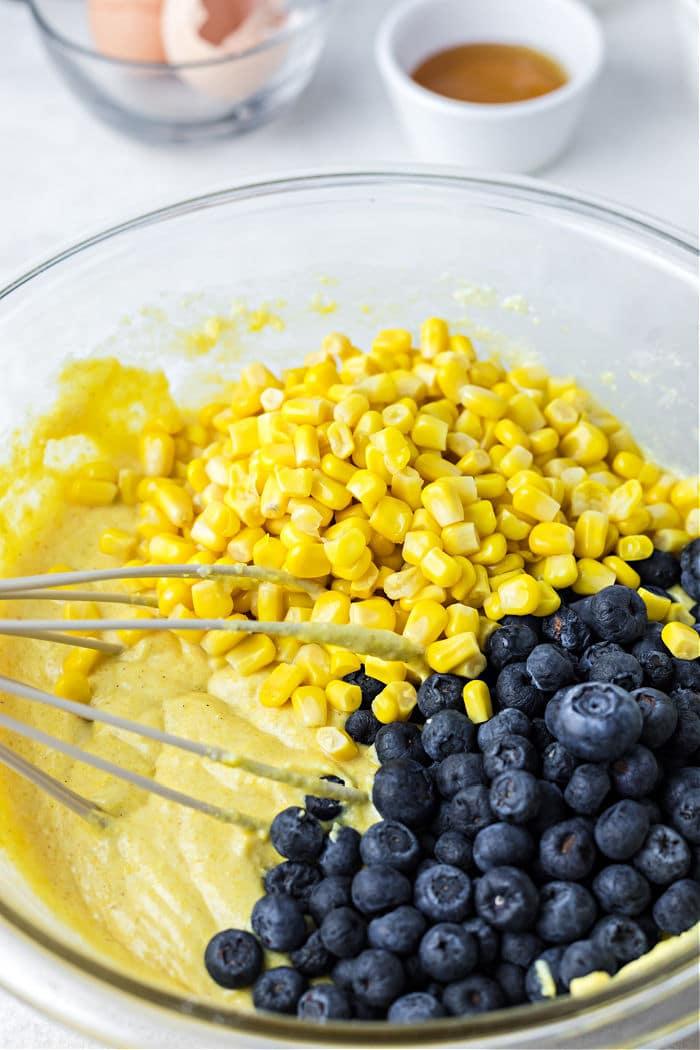 A glass bowl holds cornmeal batter adorned with corn and blueberries, whisk at the ready; eggshells and a cup linger in the background, setting the stage for a delicious jiffy cornbread baked to perfection in a cast iron skillet.