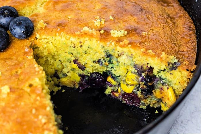 A close-up of blueberry cornbread made with Jiffy cornbread in a cast iron skillet, a slice removed to reveal its moist texture; topped with fresh blueberries.