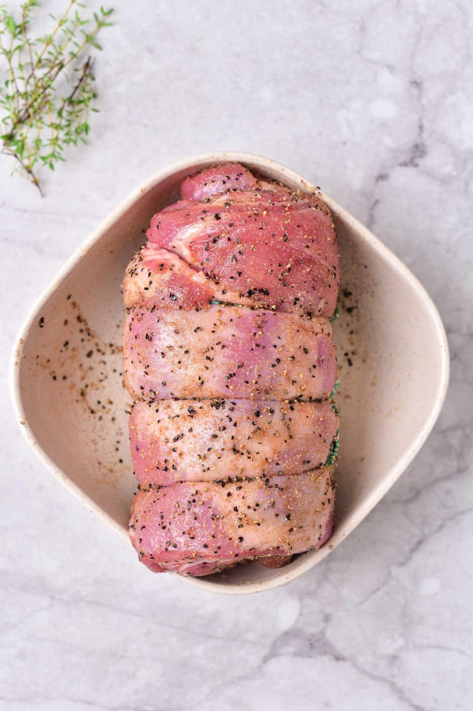 Raw seasoned rolled lamb breast in a white dish, primed for cooking, rests on a marble surface with a sprig of thyme nearby—a scene reminiscent of an exquisite lamb breast recipe.