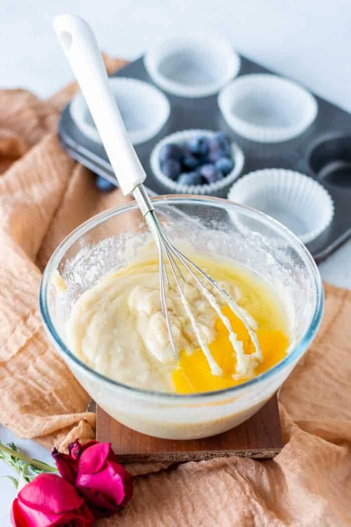 A glass bowl with muffin batter and whisk sits next to a muffin tray and rose on a beige cloth, ready for delicious frozen blueberry muffins.