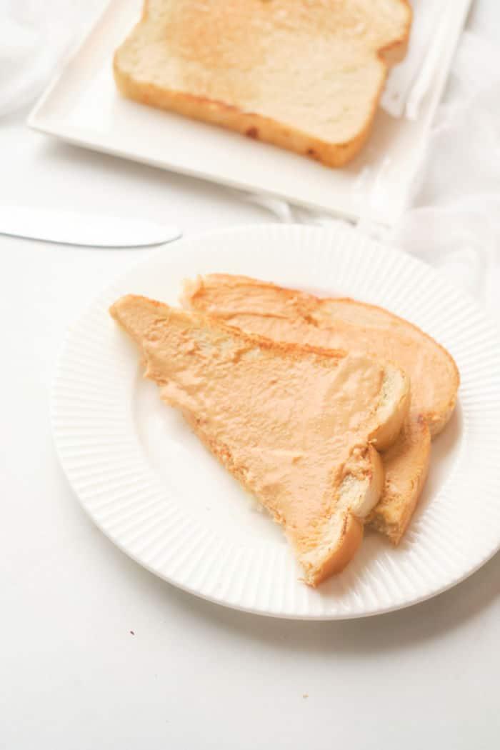 Sliced bread with peanut butter on a white plate, set on a table alongside a knife and more bread in the background, hints at the process of making toast in the oven for a warm, comforting start to your day.