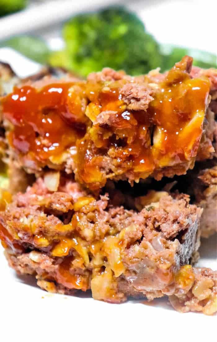 Close-up of sliced Meatloaf Without Breadcrumbs topped with ketchup glaze, with broccoli blurred in the background.
