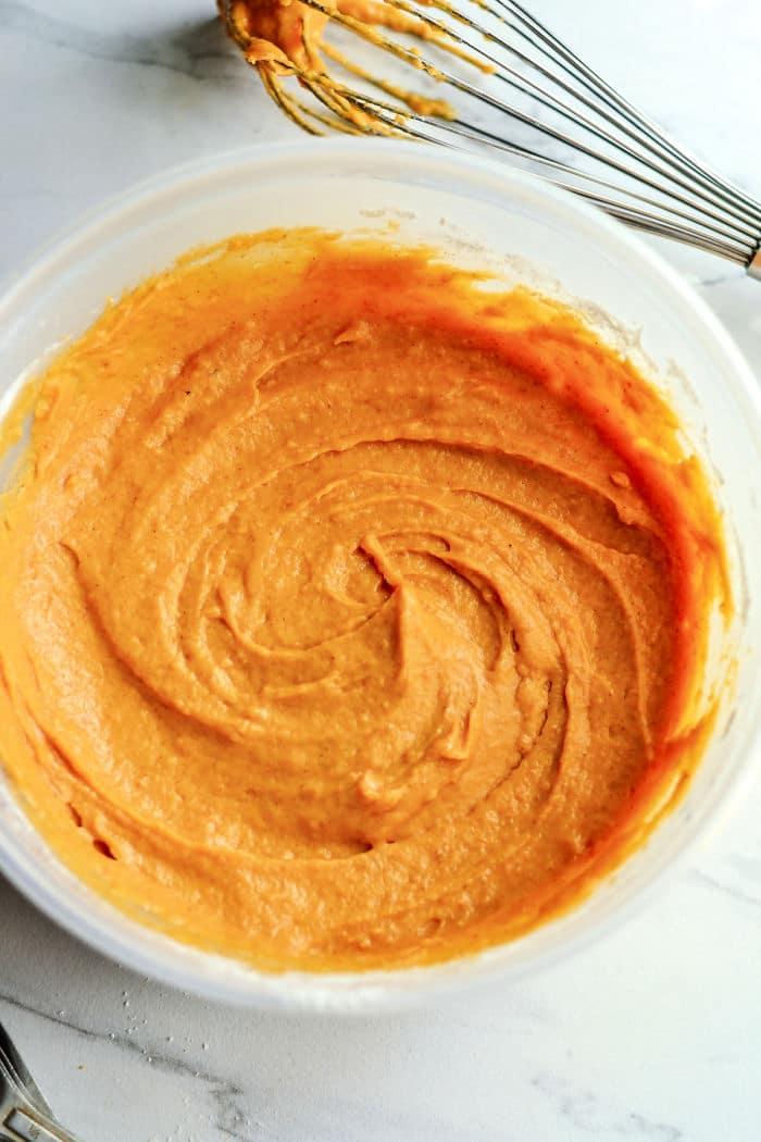 A bowl of creamy orange batter for Libbys Pumpkin Muffins with a whisk on a white marble surface.