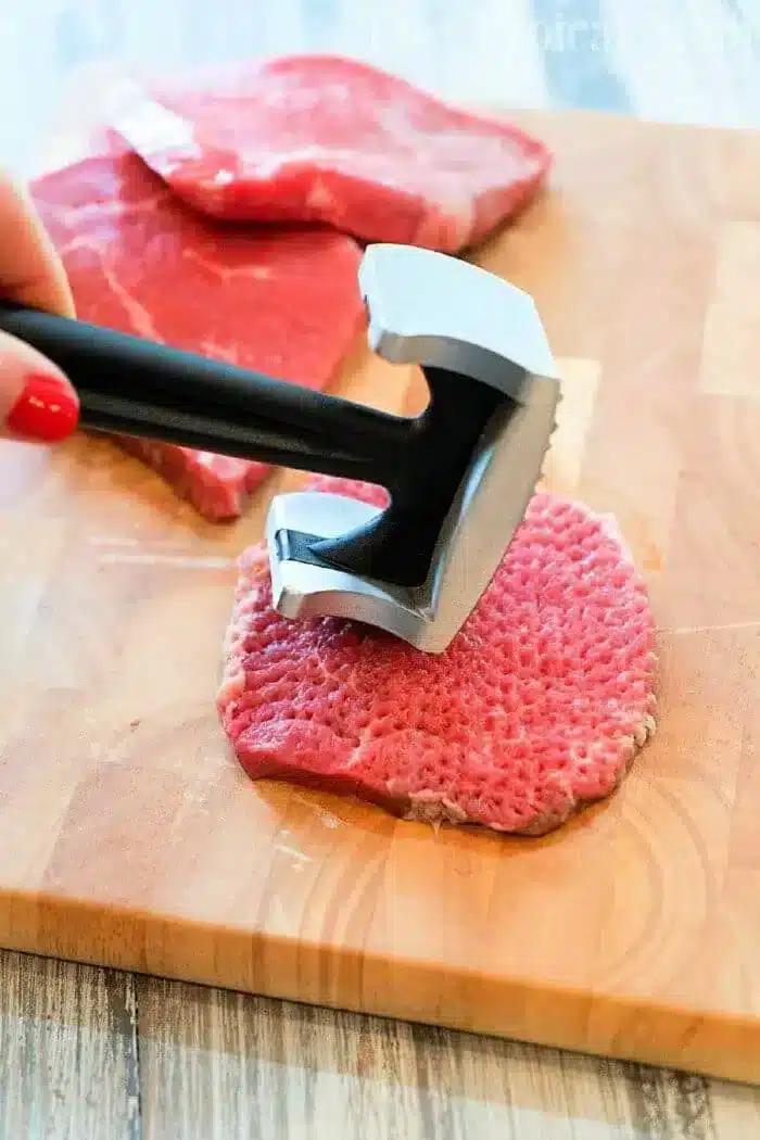 A person is expertly using a metal meat tenderizer on a piece of raw meat, preparing it for delicious minute steak recipes. The wooden cutting board holds the focus, while additional slices of meat patiently await their turn in the background.