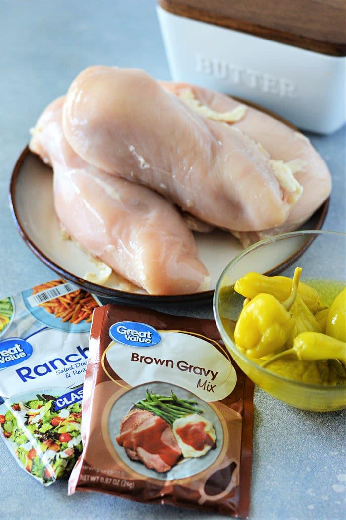 Raw chicken breasts on a plate with ranch mix, brown gravy mix, and a bowl of yellow peppers nearby—perfect for making flavorful Mississippi chicken in the Instant Pot.