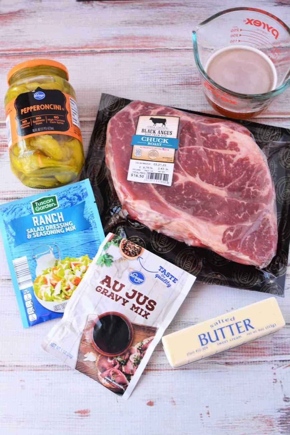Raw chuck roast, butter, pepperoncini jar, ranch mix, au jus mix, and beef broth in a measuring cup on a table—everything you need for an easy Mississippi Pot Roast Recipe.