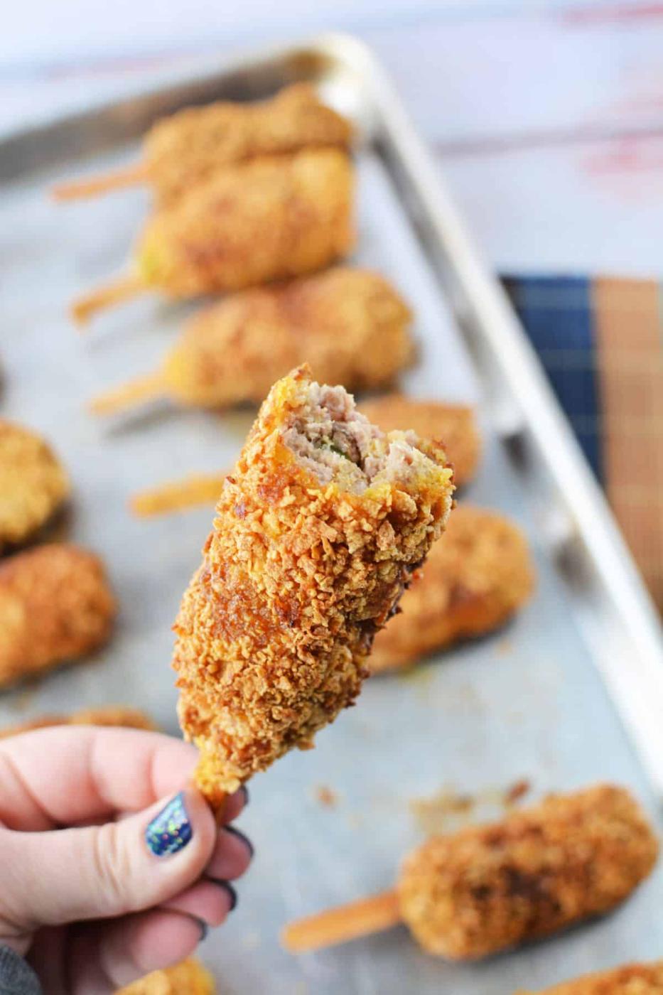 A hand holds a mock chicken leg with a bite taken out, revealing sausage inside. Nearby, mock chicken legs rest on a baking sheet, tantalizingly golden and ready to enjoy.