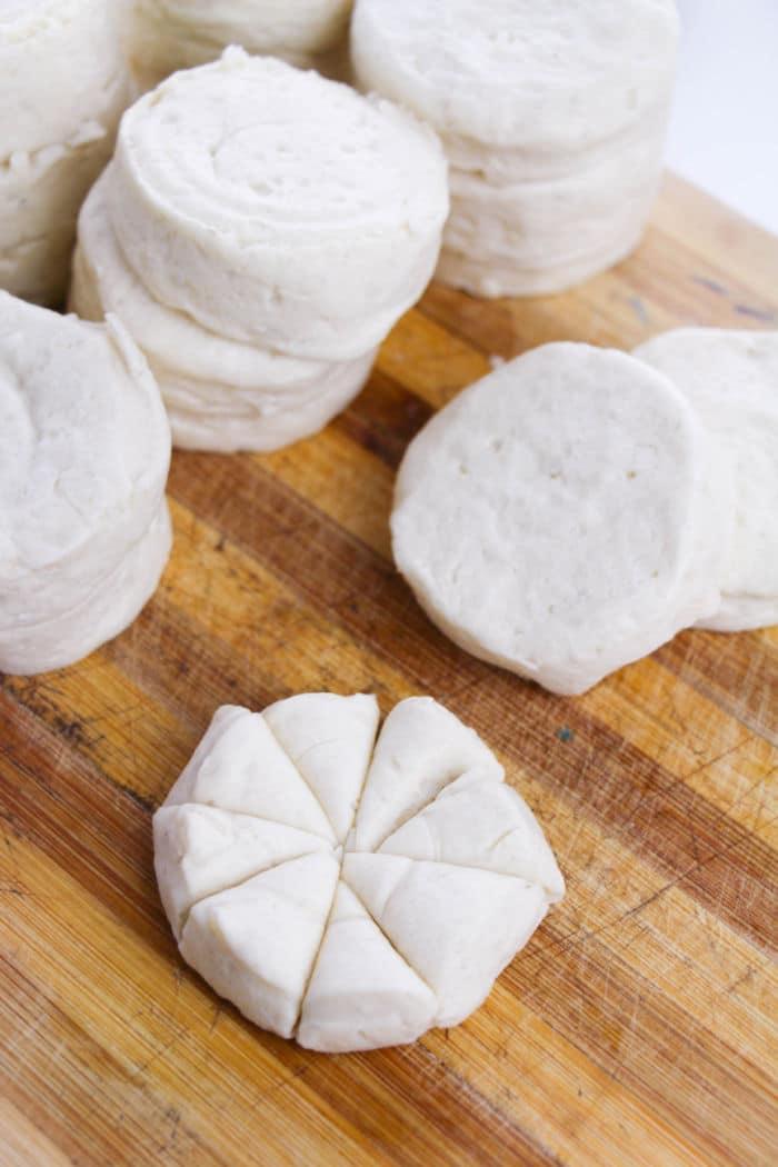 Stacks of uncooked biscuit dough, perfect for making Monkey Bread, rest on a wooden board, with one sliced into eight pieces.