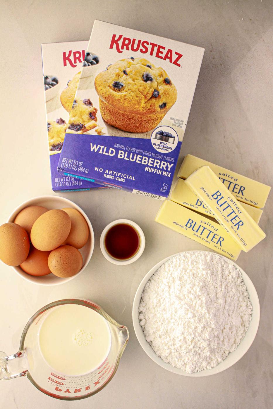 Baking ingredients: Muffin Mix Cake, eggs, butter, vanilla, milk, and powdered sugar on a countertop.