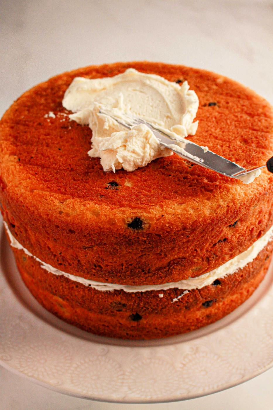 A delightful two-layered cake made with muffin mix, featuring a luscious cream filling. A knife gracefully spreads frosting on the top layer, all elegantly presented on a white plate.