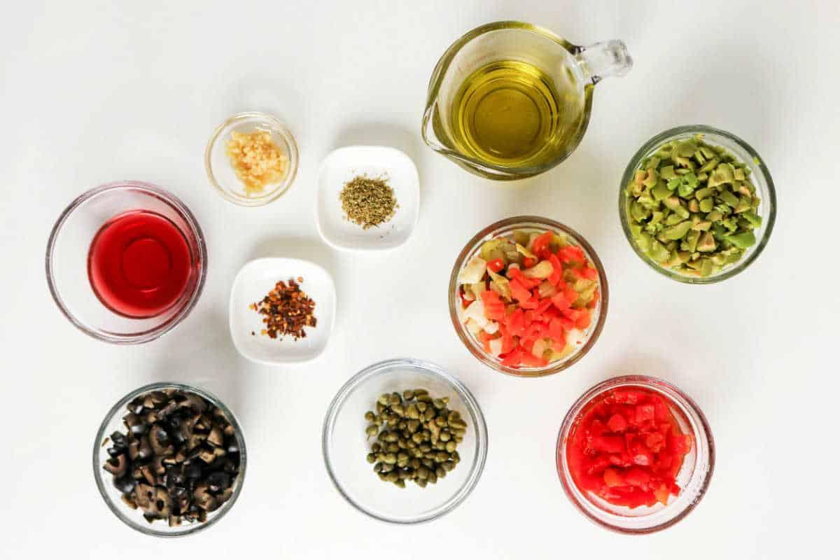 Various small bowls with chopped vegetables, olives, capers, spices, oil, and vinegar arranged on a white surface—perfect ingredients for a classic Muffuletta recipe or homemade olive salad.