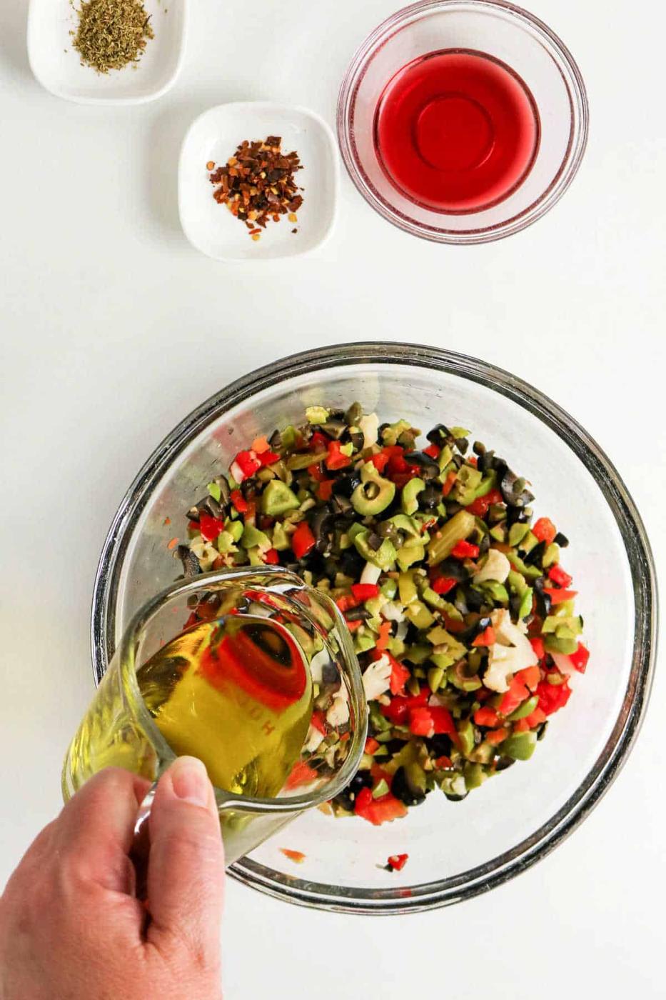 A hand pours olive oil into a bowl of chopped vegetables for a classic Olive Salad, with spices and red vinegar nearby—perfect for an authentic Muffuletta recipe.