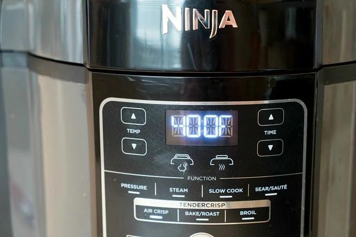 Close-up of a black Ninja multi-cooker control panel equipped with Ninja Foodi accessories. The digital display reads "00:00." Buttons for different functions such as pressure, steam, slow cook, sear/sauté, and more are visible.