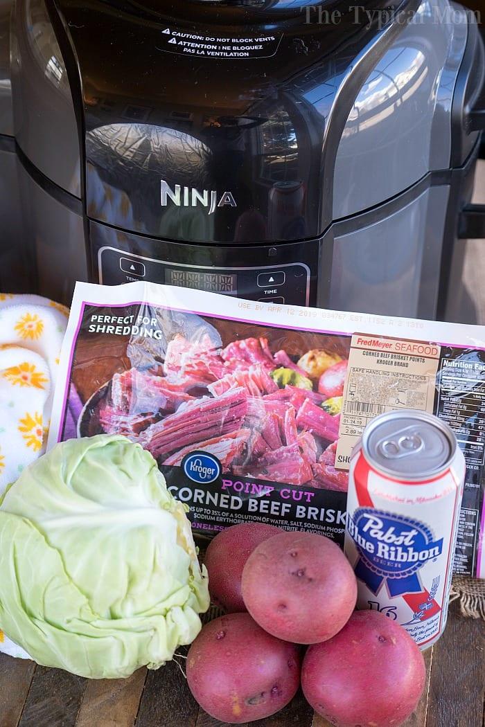 On a wooden table sits a tempting array: cabbage, red potatoes, canned beer, and a corned beef brisket package. Perfect for creating the ultimate Ninja Foodi corned beef and cabbage feast.
