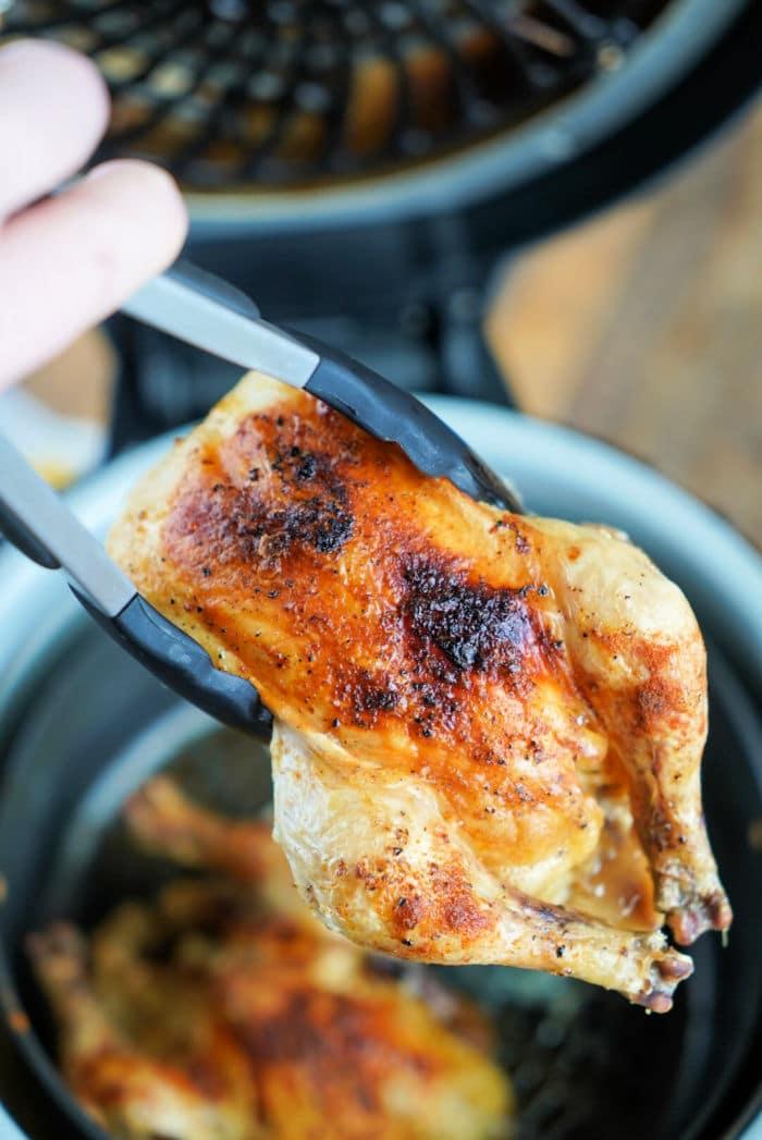 Tongs holding a piece of seasoned, cooked cornish hen with crispy skin above a Ninja Foodi basket, perfect for achieving that ideal crispy texture.
