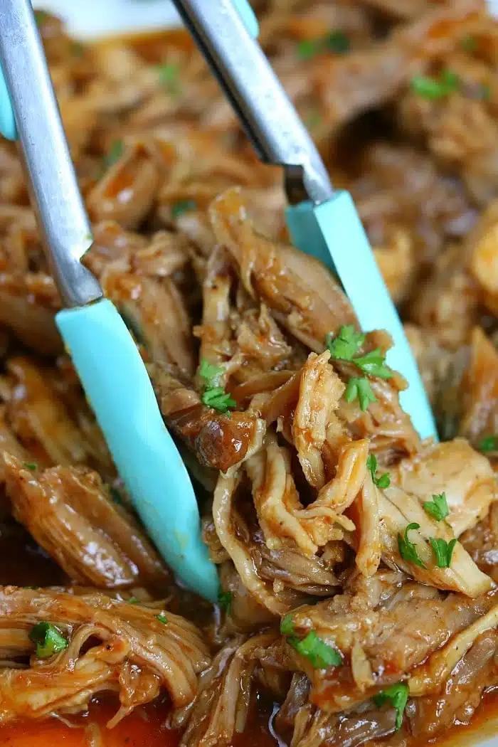 Tongs holding shredded, saucy pulled pork garnished with chopped parsley—perfect for trying out new ninja pressure cooker recipes.