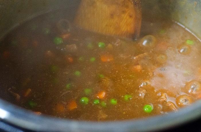 Stew with peas and carrots simmering in a Ninja Foodi, stirred with a wooden spoon.