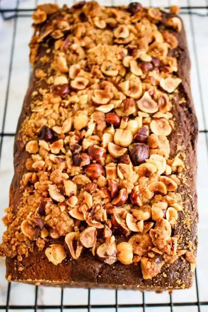 A loaf nutella bread topped with a crumbly mix of chopped nuts, including hazelnuts, reminiscent of Nutella bread, sits on a cooling rack. The golden-brown cake has a textured top with scattered pieces of nuts and crumbs visible.