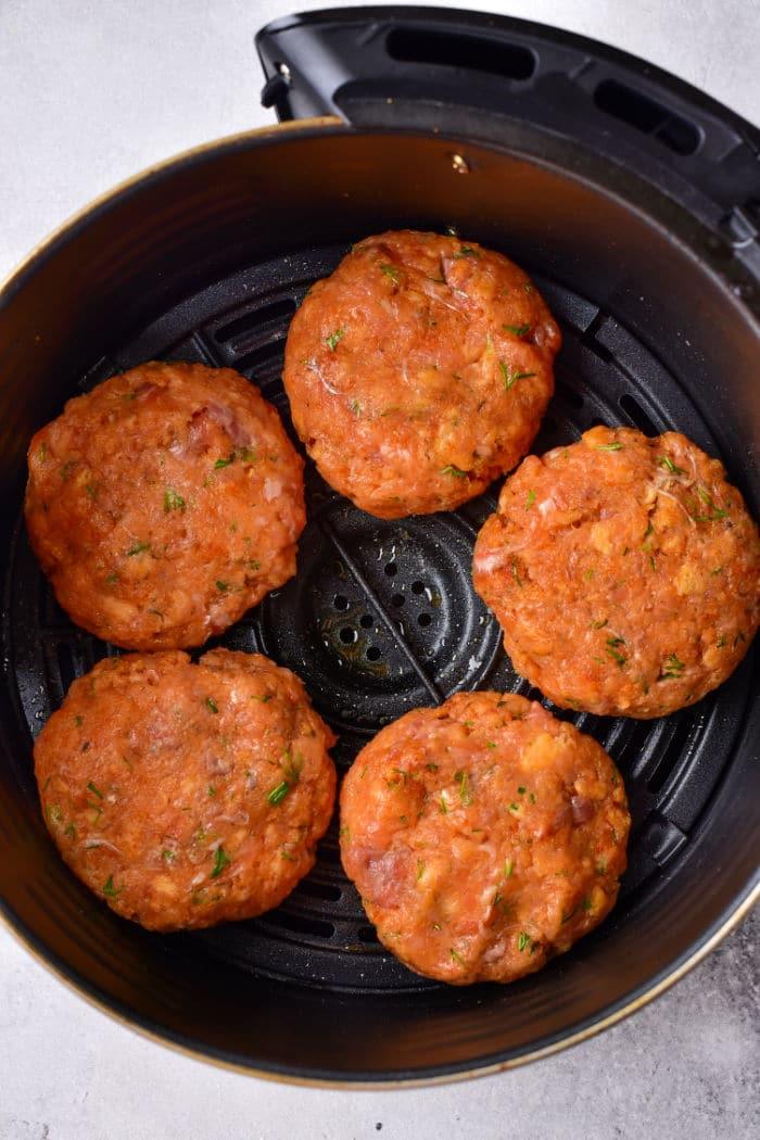 Five raw salmon patties seasoned with herbs sit in an air fryer basket, ready for a modern twist on cooking. These aren't your Old Fashioned Salmon Patties, but they're sure to awaken nostalgic flavors with every bite.