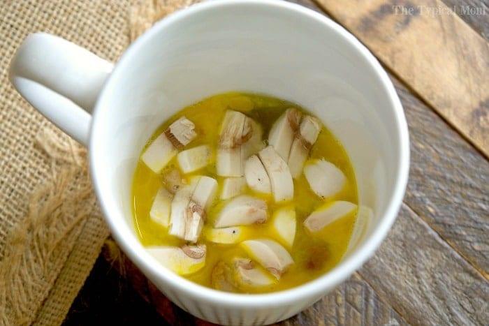 A white mug filled with a eggs and sliced mushrooms, reminiscent of an omelette in a mug, rests on a wooden surface.