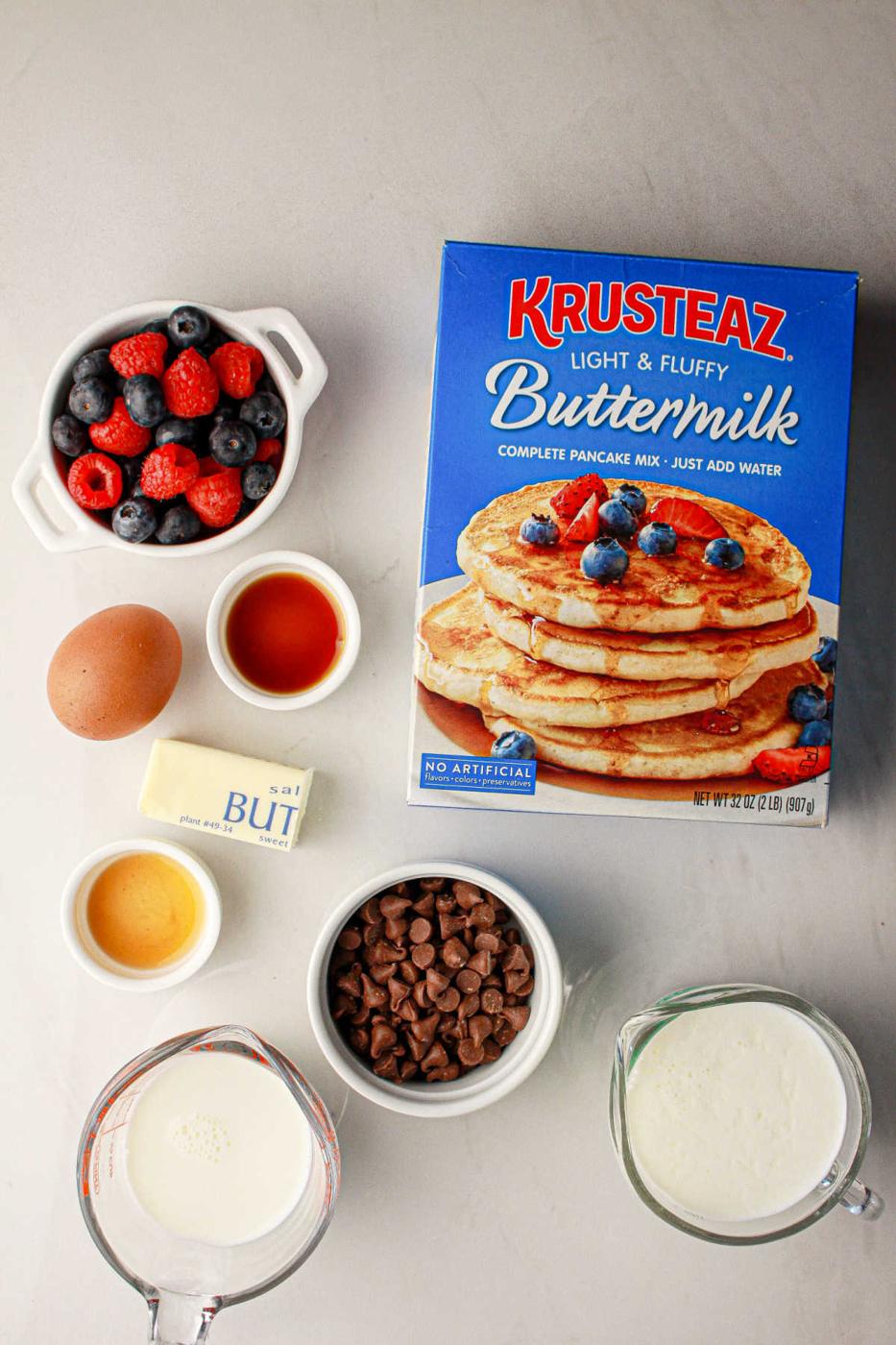 Box of pancake mix, berries, chocolate chips, butter, milk, egg, vanilla, and syrup on a gray surface—everything you need for delicious oven baked pancakes with pancake mix.