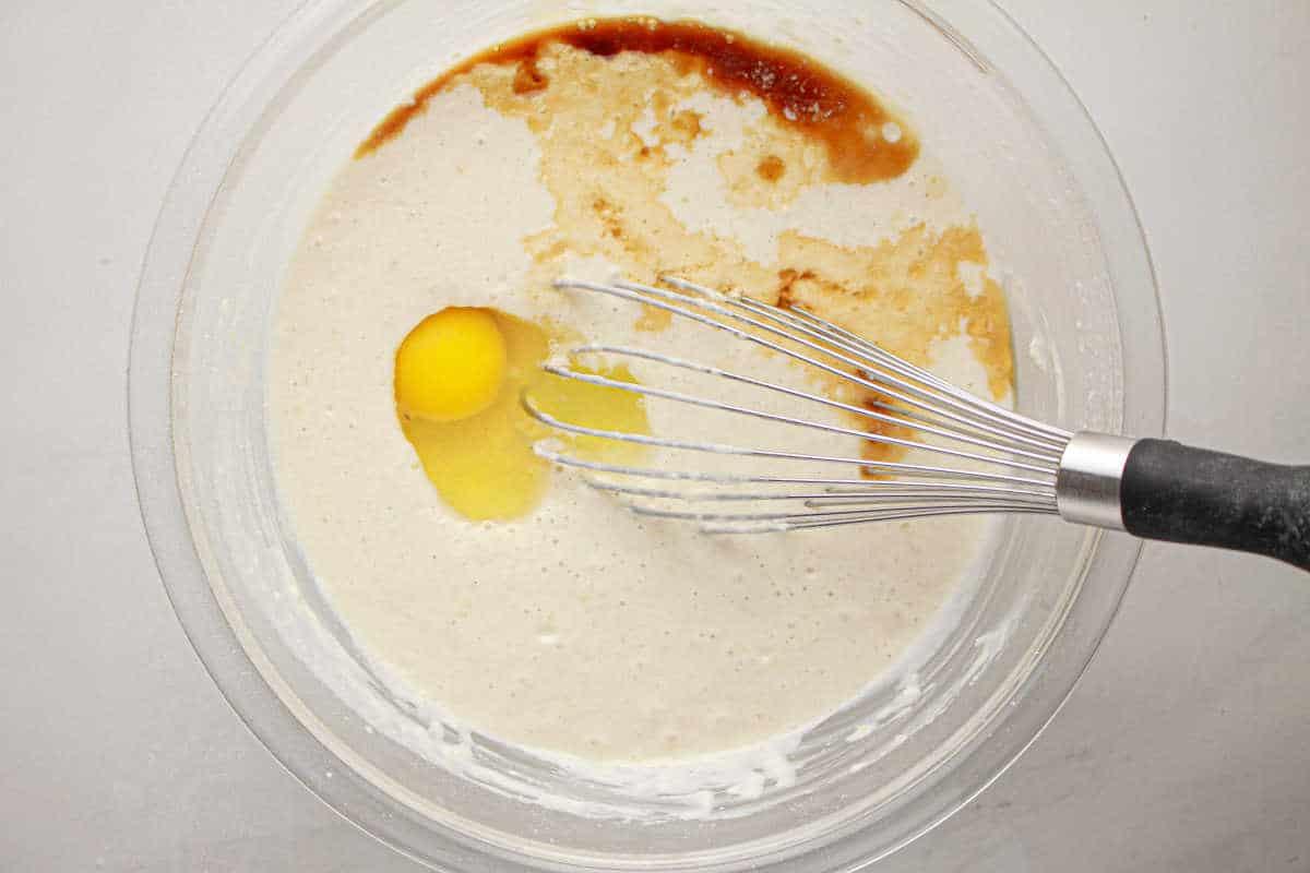 A whisk is in a bowl with batter, an egg, vanilla, and other ingredients being mixed together for oven baked pancakes with pancake mix.