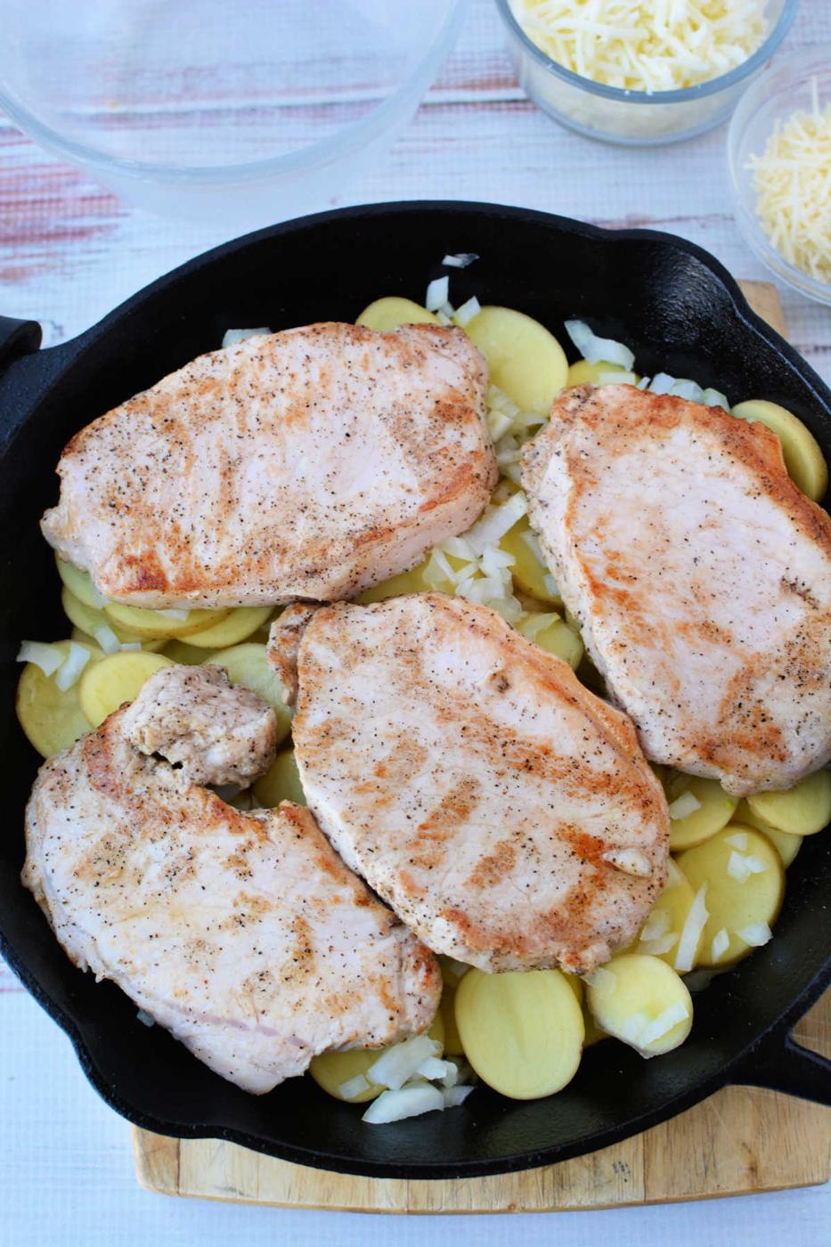 Four seasoned oven baked pork chops rest on sliced potatoes and chopped onions in a black skillet, with cheese in the background.