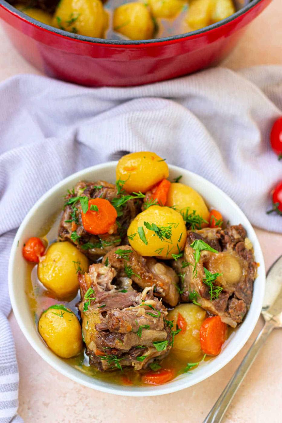 A bowl of ox tail soup with potatoes, meat, and carrots garnished with herbs, alongside a red pot and wooden spoon.