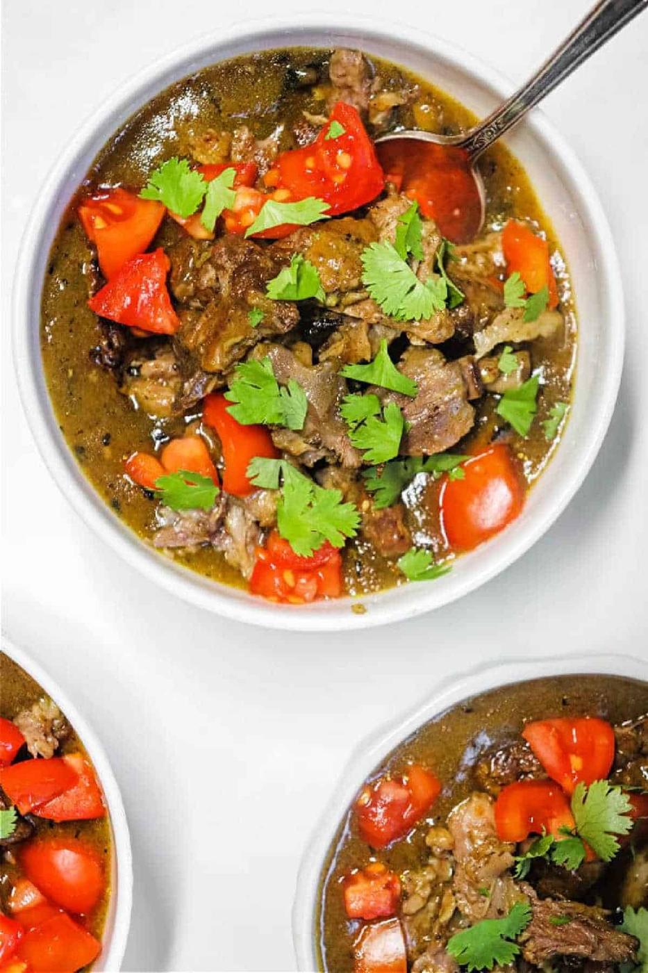 A bowl of hearty oxtail curry stew topped with chopped tomatoes and fresh cilantro, with a spoon in the bowl.