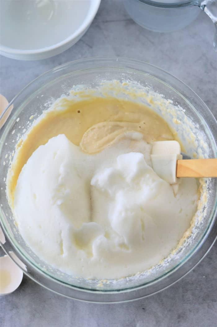 A glass bowl containing whipped egg whites being gently folded in using a spatula. The bowl rests on a gray countertop, with another bowl and kitchen items visible in the background.