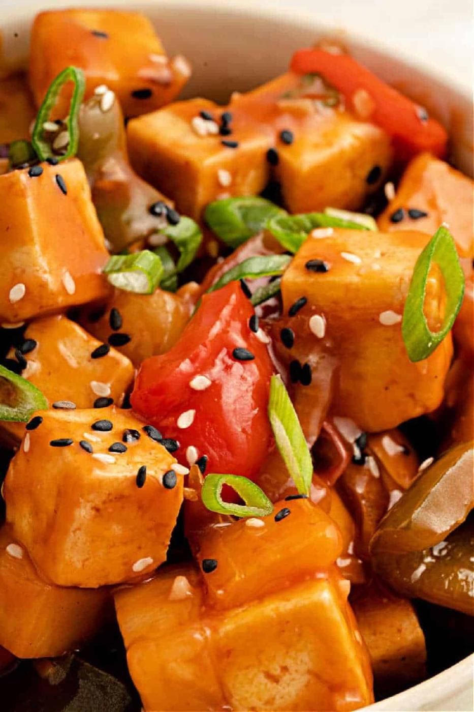 Close-up of tofu cubes in Copycat Panda Express Sweet and Sour Sauce, with bell peppers, sesame seeds, and sliced green onions.