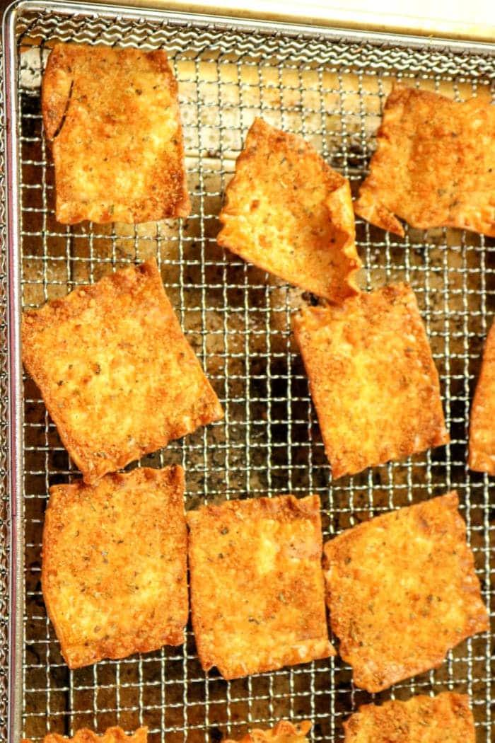 A close-up view of golden-brown, crispy pasta chips cooling on a metal wire rack. These uniformly square bites, with some displaying a jagged edge on one side, were crafted in an air fryer. The wire rack is situated on a light surface.