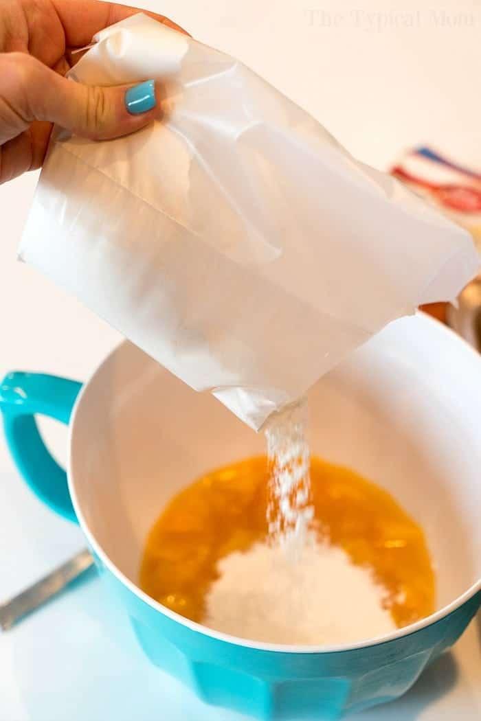 A hand pours white powder from a peach angel food cake packet into a blue bowl containing beaten eggs.