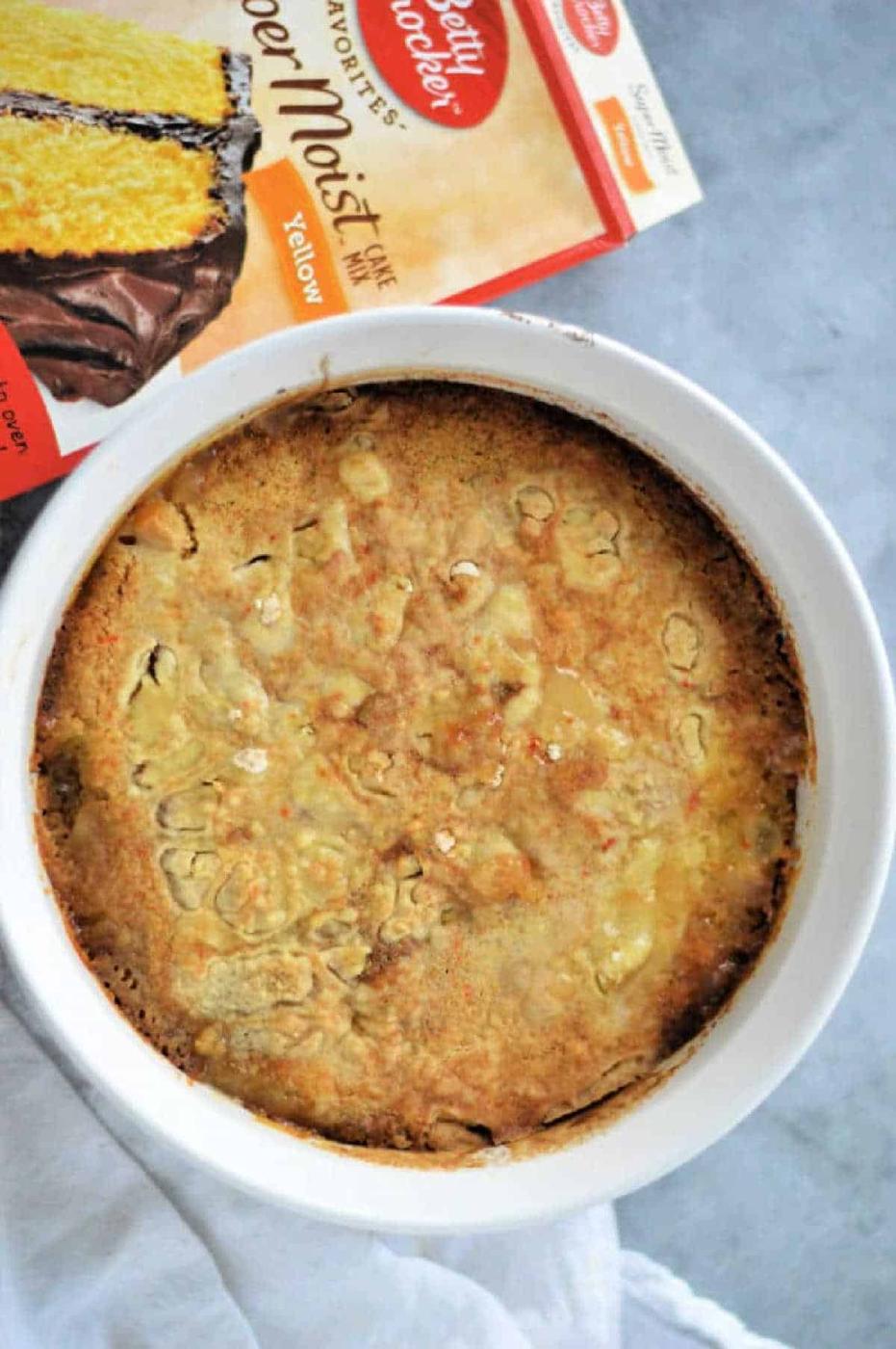 A peach cobbler with cake mix is baked in a white dish, sitting next to a Betty Crocker yellow cake mix box on a gray surface.