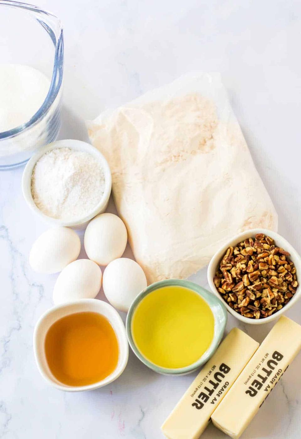 The table is set for a Pecan Bundt Cake masterpiece with bowls and packages of flour, sugar, eggs, pecans, butter, oil, and vanilla extract.