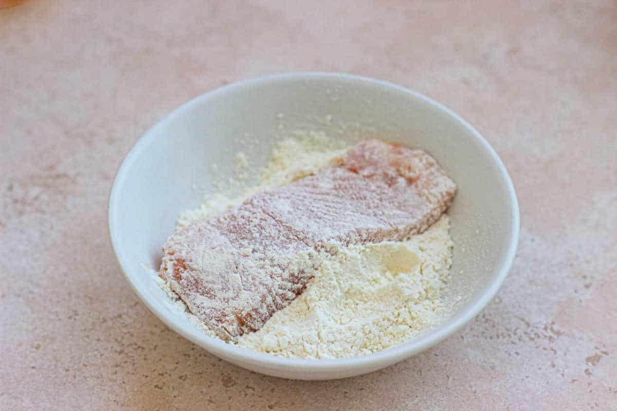Raw trout coated in flour sits in a white bowl on a beige countertop, reminiscent of preparing pecan-crusted trout.