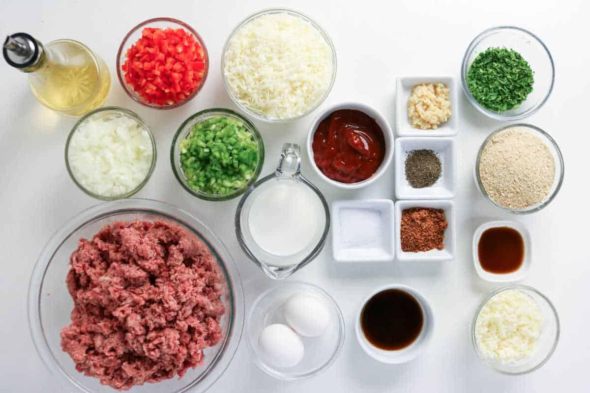 Ground meat, eggs, vegetables, seasonings, cheese, breadcrumbs, milk, and sauces arranged on a white surface—everything you need to craft a delicious Philly Cheesesteak Meatloaf bursting with classic flavors.