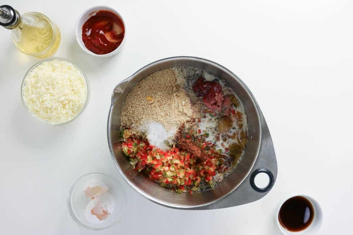 Metal bowl with eggs, breadcrumbs, diced peppers, and seasonings—perfect for mixing up a delicious Philly Cheesesteak Meatloaf. Cheese, ketchup, and sauces are arranged in small bowls nearby to enhance the classic flavors of this meatloaf-inspired dish.