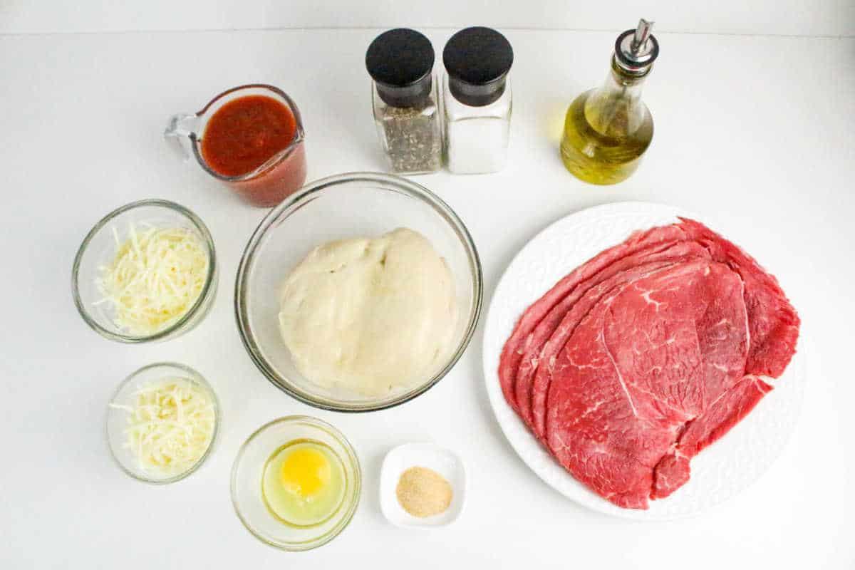 Philly Cheesesteak Stromboli ingredients: pizza dough, raw steak slices, cheese, egg, marinara sauce, oil, salt, pepper, and seasoning arranged on a white surface.