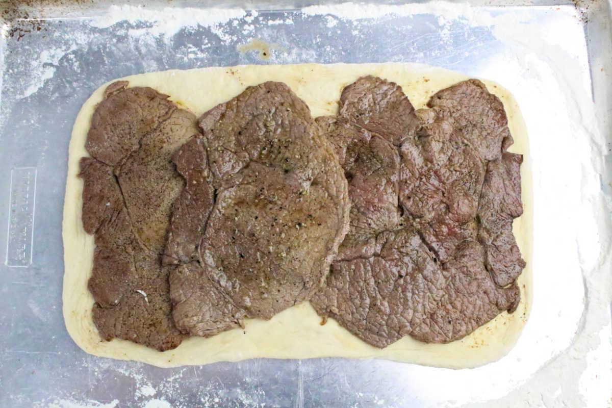 Rectangular dough topped with several overlapping slices of cooked beef, ready to be transformed into a delicious Philly Cheesesteak Stromboli on a floured baking sheet.