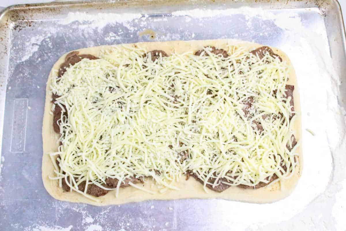 Rectangular pizza dough topped with meat slices and shredded cheese on a floured baking sheet, perfect for making Philly Cheesesteak Stromboli.