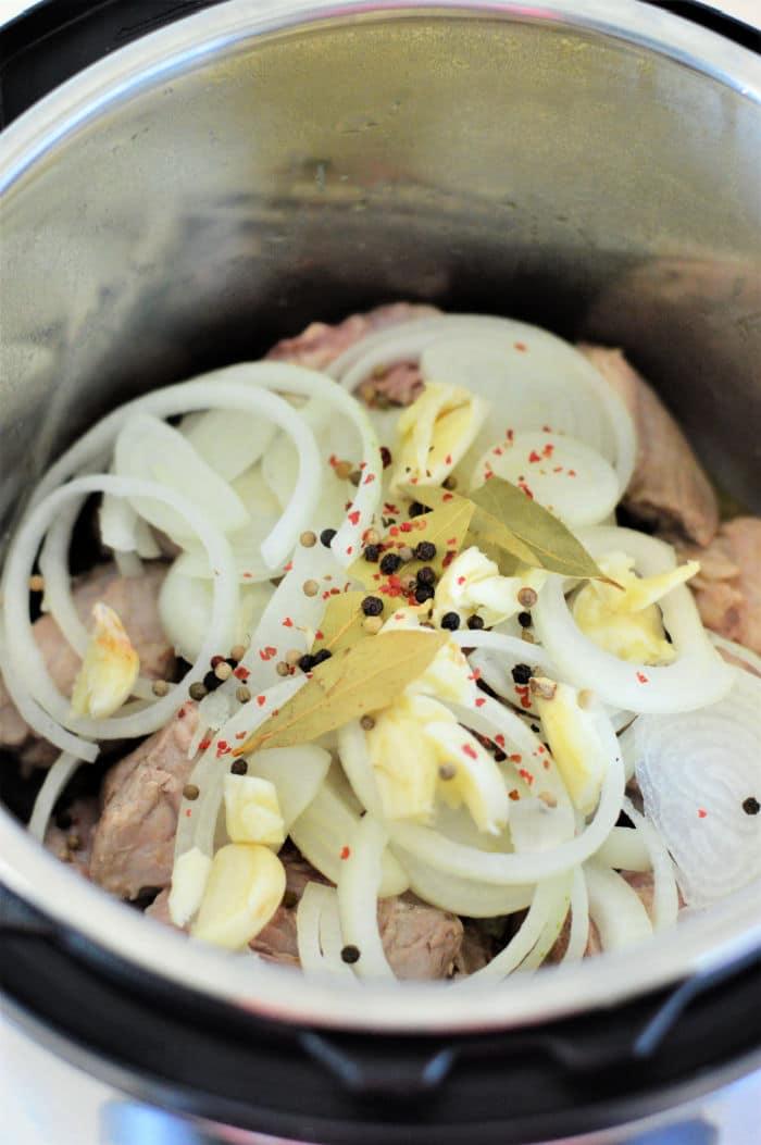 Sliced onions, garlic, bay leaves, and peppercorns on top of meat in this easy pork adobo Instant Pot recipe.