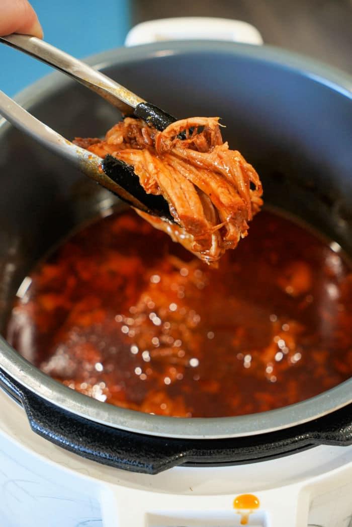 Tongs holding shredded pork butt over an Instant Pot filled with red sauce.